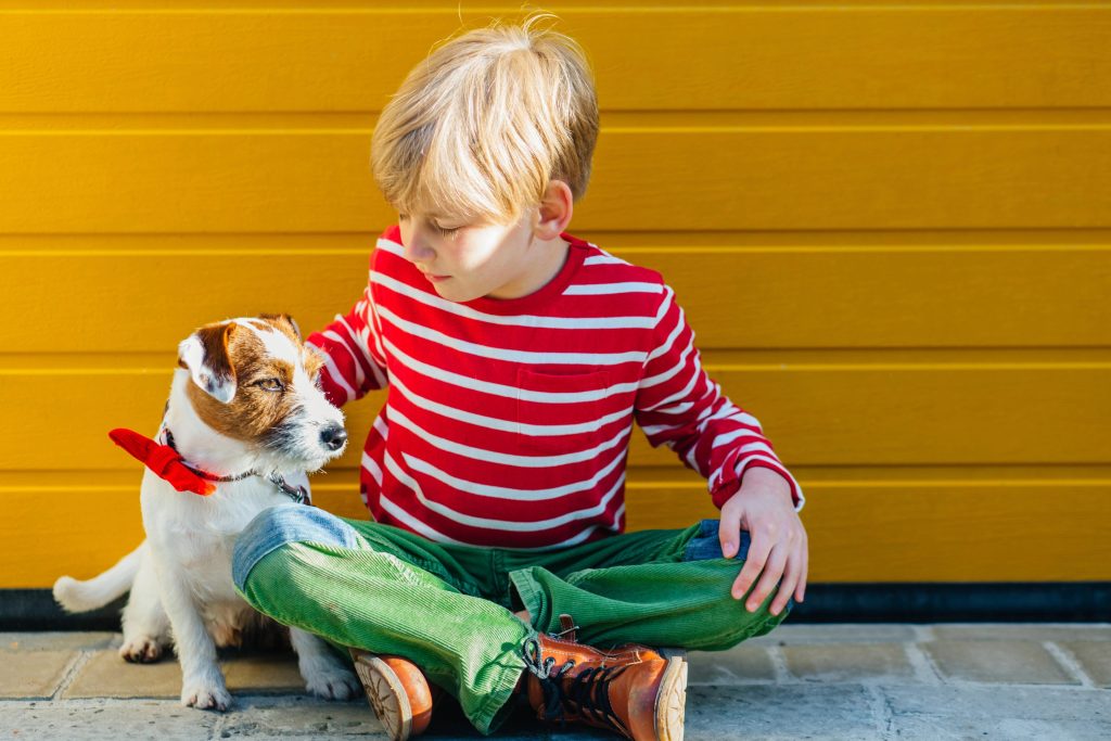 A young boy sat outside a yellow garage door, stroking a small dog.